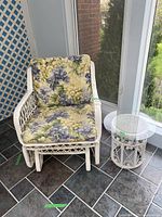 White wicker armchair with floral cushions and matching white wicker round side table with glass top on tiled floor in enclosed porch area.
