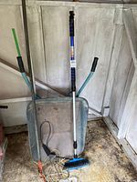 Wheelbarrow with blue handles and metal tray, push broom leaning inside, visible dust and dirt on the floor.