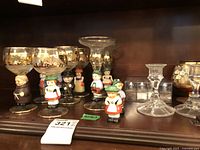 Photo of Goebel goblets and ceramic figurines displayed on a wooden shelf, showing gold trim and multiple glass items.