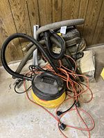 Photo showing two yellow and black wet/dry vacuums with hoses and tangled orange electrical cords on a concrete floor against a wood-paneled wall.