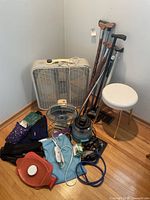 Photo showing white round stool, box fan, standing fan, wooden and metal crutches, and a collection of health and therapy items on floor.