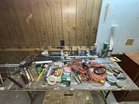 Wide view of assorted tools on the table including extension cords, hand saws, hammers, paint brushes, rolls of tape, and miscellaneous electrical items.