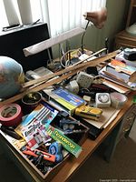 Top view of cluttered office desk with globe, desk lamps, utility tools, batteries, stationery and miscellaneous accessories scattered and stacked.