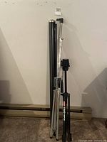 Folded Optex T-300 projector screen in a black metal casing, standing next to its tripod stand with black legs and silver adjustment mechanisms on a carpeted floor.