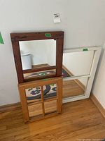 Three wooden framed mirrors leaning against a wall on a hardwood floor: dark stained rectangular frame, white large square frame, light wood small rectangular frame.