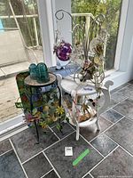 Overview photo of two small side tables, one black with floral glass inserts and one white, with several hanging stained glass decorations and vintage green glass insulators on the black table.