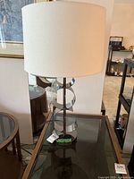 Full view of the table lamp showing silver sculptural metal base and white fabric shade on glass-topped table