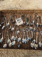 Photo showing a wide array of assorted souvenir spoons laid out on woven surface, featuring metal and enamel decorative handles with various minor wear.