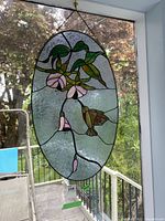 Hanging oval stained glass panel with colorful hummingbird and flowers design visible against window light.