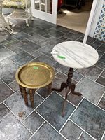 Photo showing both decorative tables on tiled floor, brass small table with ornate top and wooden legs, larger white marble-like table with wooden base