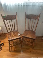 Pair of traditional wooden chairs with carved floral backrests and spindle detailing, set on hardwood flooring beneath a window with vertical blinds.