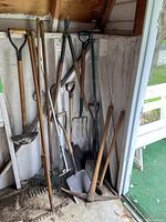 Photo showing various garden tools including rakes, shovels, pickaxe, and pitchfork all leaning against a wall inside a shed area.
