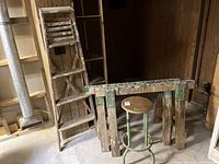 Photo of the wooden ladder folded and leaning against the wall next to the sawhorse and stool, showing wooden slats and hinges.