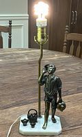 Table lamp with bronze water boy sculpture, brass lamp fixture, and marble base, shown lit with bulb in socket. Three small bronze vessels and power cord visible on base.