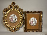 Both vintage framed Italian Baroque style plaques shown side by side against a neutral background, front view. Ornate gold frames and oval 3D relief plaques featuring a Victorian man and woman seated in a garden scene.