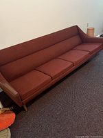 Full view of long brick red wool upholstered sofa with teak legs and armrest accents in living room setting.