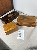 Three wooden boxes arranged on a cloth surface showing top and side views. The items include a dark brown box with a metal plaque, a decorative inlaid wood box, and a burl wood box.