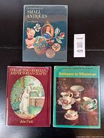 Three hardcover antique reference books placed on a dark surface showing covers: one about small antiques, another about Georgian and Victorian crafts, and one on antiques in miniature.