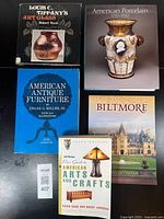 Five American antique reference books shown front-facing on a black surface, titles visible, arranged in two rows: Louis C. Tiffany's Art Glass, American Porcelain 1770-1920, American Antique Furniture, Official Price Guide to American Arts and Crafts, and Biltmore.