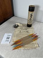 Photo showing antique 456 Pick Up Sticks game pieces including wooden sticks with colored tips, metal scoop, instruction sheet, and original cylindrical tube container with cap.