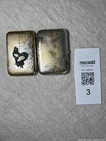 Open view of metal case showing crane design etched inside surfaces, black enamel accent on perched crane, visible wear and tarnish.