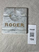 Top view of square marble ashtray with carved grooves, raised circular ashwell, and gilt letters spelling 'Roger'.