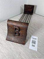 Close-up of one end of the extendable book rack showing dark oak bookend with carved letter B monogram and brass rods.
