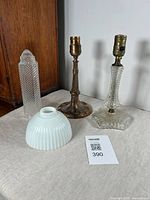 Photo showing two lamp bases (bronzed cast spelter and pressed glass with brass sockets) along with milk glass and clear glass shades on a table.