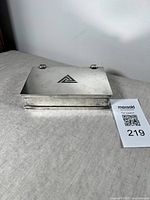 Front view of the silver plated hinged brass box with visible triangular design on the lid and two metal hinges at back.