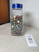 Clear plastic jar filled with colorful vintage clay marbles from a side angle, showing condition and quantity.