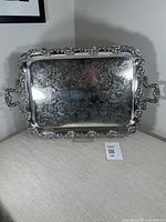Front view of large rectangular silver plate tray with ornate floral handles and detailed engraved pattern on surface placed on a beige cloth.