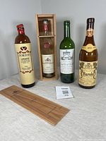 Four vintage bottles including Duboeuf Apéritif, Montesquieu Armagnac in wooden box, Grand Vin De La Croix white wine, and 1979 Barolo wine on grey surface with wooden lid and lot tag.