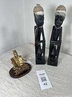 Photo showing two tall Indonesian hardwood figures standing side by side with fiber headdresses and an adjacent smaller cast metal musician figure seated on a hardwood base.