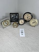 Front view of all six vintage alarm clocks in a group on a beige cloth surface, showing varying sizes and colors