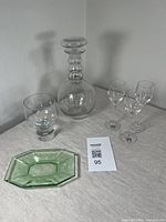 Full view of the lot showing clear glass decanter with stopper, three clear glasses, one tumbler glass and a green glass tray on a table.