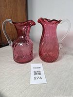 Pair of cranberry glass pitchers with clear handles displayed on neutral fabric surface showing overall size and color.