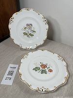 Two antique cabinet plates displayed on stands, showing floral motifs and gold scalloped rims