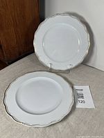 Two white scalloped edge ironstone plates with gold gilt trim, displayed on plain stands against neutral background. One plate is more heavily crazed with a visible crack, the other in better condition with minor gilt losses.