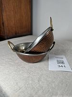 Two nested bowls showing hammered copper outer surface and brass handles, placed on a beige cloth.