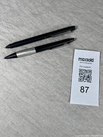 Top-down view of two Lamy pens on grey fabric surface with MaxSold lot tag showing lot number 87