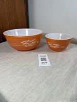 Pair of two vintage Pyrex mixing bowls with orange exterior and white interior, decorated with wheat motif.