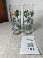 Two tall clear vintage glasses with repeating green cactus wearing headphones graphics and Seagram's Gin & Sonic text on a white cloth background with MaxSold paper.