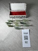 Full lot of 10 jade fish-shaped chopstick rests arranged in two rows in front of a decorative fabric-covered box with red lining.