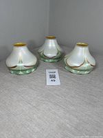 Three opaque iridescent white floriform art glass shades with ruffled rims. Green pulled feather design with gold tips visible. Arranged on a light cloth surface.