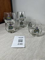 Six clear glass tumblers with RCMP insignia arranged spaced out on a beige cloth surface.
