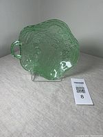 Green uranium glass bowl with handle, showing embossed cherry and leaf patterns and textured background, photographed on a stand against a white background.