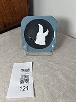 Front view of the square plaque showing blue background, black circular center, and white goose motif