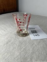 Side view of a clear glass tumbler with red nautical flag and pennant designs, standing on a fabric surface next to a lot number card.