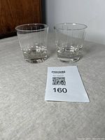 Pair of vintage glass tumblers with vertical cut lines shown side by side on a beige fabric surface with wooden furniture in background.