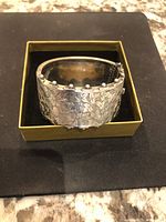 Wide sterling silver bangle bracelet with intricate engraved ivy leaf pattern and beaded edges, placed in a yellow box on a black surface.